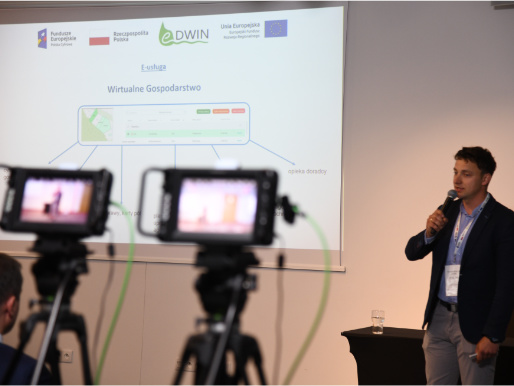 Na pierwszym planie widoczne są dwie kamery nagrywające widocznego z tyłu Łukasza Kuleczkę, doradcę WODR, który stoi z mikrofonem obok widocznej prezentacji.