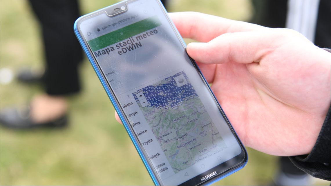 Zbliżenie na dłoń, w której trzymany jest telefon komórkowy. Na ekranie telefonu widać mapę z lokalizacją stacji meteorologicznych.