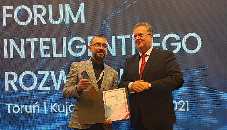 Maciej Zacharczuk (po lewej) stoi obok mężczyzny w średnim wieku i trzyma nagrodę z dyplomem. Na niebieskim tle z tyłu jest napis "Forum Inteligentnego Rozwoju".