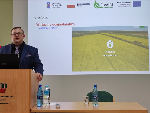 Wnętrze pomieszczenia. Michał Głombicki, młody mężczyzna, stoi w marynarce przed rzutnikiem, na którym widoczna jest prezentacja o wirtualnym gospodarstwie. Przed nim jest mównica, na której stoi laptop.