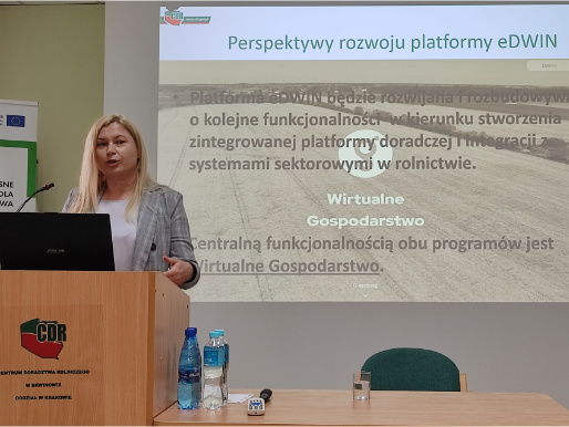 Wnętrze pomieszczenia. Anna Pietruczuk, młoda kobieta, stoi w marynarce przed rzutnikiem, na którym widoczna jest prezentacja o Wirtualnym gospodarstwie. Przed nią jest mównica, na której stoi laptop.