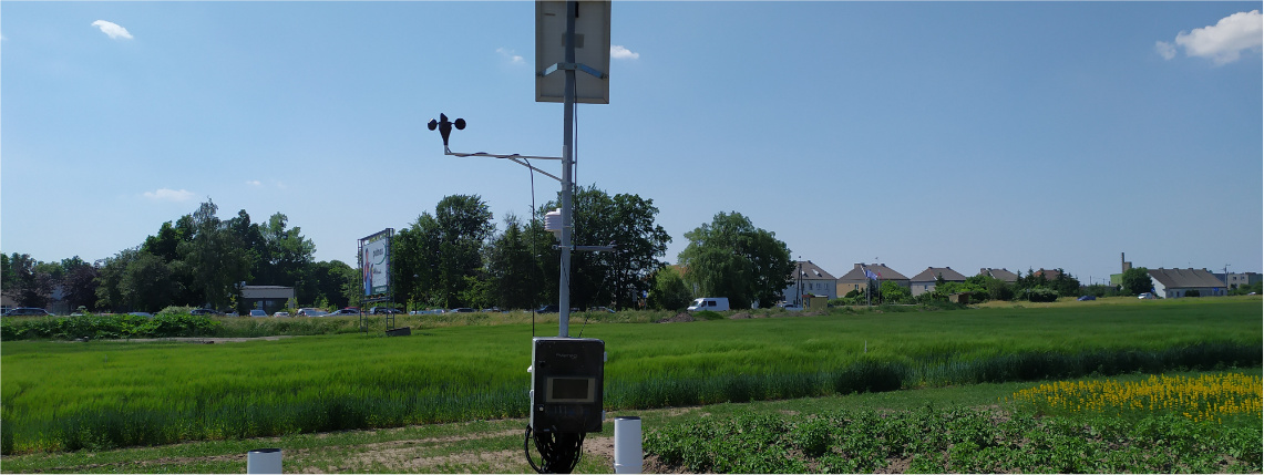 Słoneczny dzień. Na polu stoi stacja meteorologiczna, która zbiera dane meteorologiczne. W tle widać uprawy i drzewa.