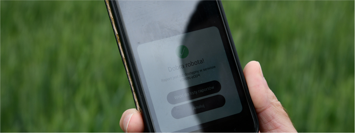 Zbliżenie na ekran telefonu, który znajduje się w dłoni. Na ekranie widać panel z napisem "Dobra robota! Raport jest już dostępny w serwisie eDWIN eODR". Poniżej są przyciski "wróć do listy raportów" oraz "anuluj".