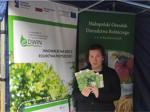 Stoisko eDWIN podczas Giełdy rolniczej w Łącku Młoda kobieta w czarnej bluzie trzyma ulotki promujące eDWIN. Za nią widać rollup promujący platformę eDWIN oraz rollup Małopolskiego Ośrodka Doradztwa Rolniczego.