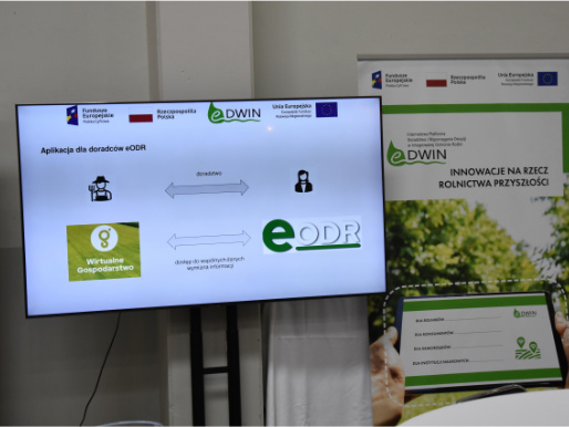 Stoisko eDWIN na Targach Bio Green Future Wnętrze hali targowej. Po lewej stronie stoi telewizor, na którym wyświetlone są informacje o platformie eDWIN. Po prawej stronie stoi rollup promujący platformę eDWIN.