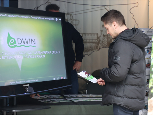Stoisko eDWIN Wiosennych Targach Ogrodniczych w Olsztynie Młody mężczyzna w czarnej kurtce stoi i przygląda się ulotce eDWIN, którą trzyma w ręku. Obok niego jest ekran telewizora, na którym widać logo eDWIN.